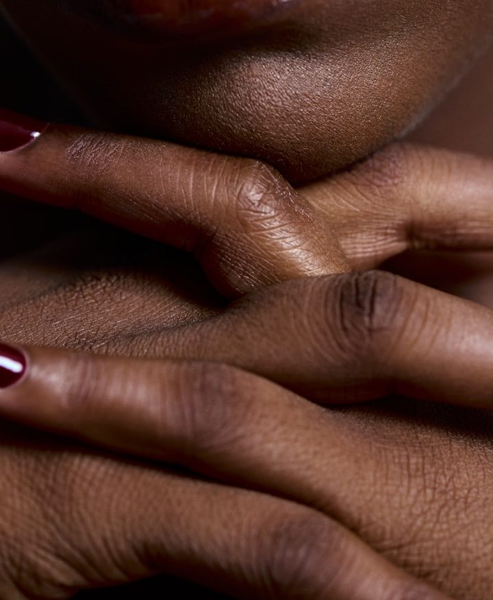 Detailaufnahme von gekreuzten Händen mit rotem Nagellack auf dunkelbrauner Haut.